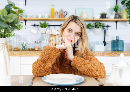 Jeune femme assise à la table de cuisine devant une assiette vide. Arrière-plan flou. Banque D'Images