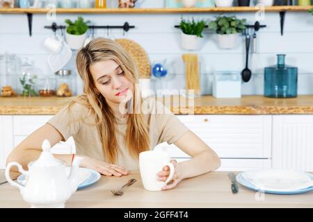 Une jeune femme s'assoit à la table de cuisine avec une tasse sur la main. Arrière-plan flou. Banque D'Images