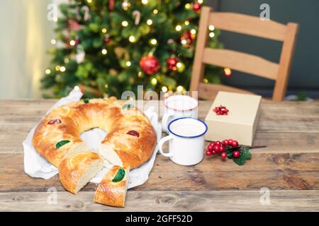 Roscón de Reyes (typique espagnol sucré) coupé, sur une table en bois et un arbre de Noël avec des lumières en arrière-plan. Bonbons et desserts de Noël. Banque D'Images