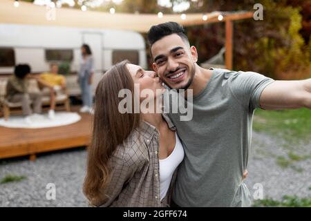 Couple multiracial aimant prenant selfie près de motorhome, embrassant et embrassant, se reposant avec leurs amis à l'extérieur Banque D'Images