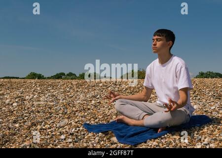 Un adolescent de race blanche méditant sur une plage pierreuse avec ses mains en position Shuni Banque D'Images