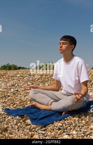 Un adolescent de race blanche méditant sur une plage pierreuse avec ses mains en position Shuni Banque D'Images