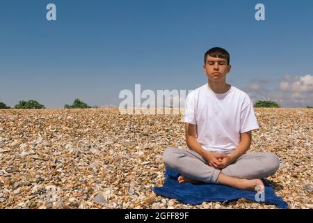 Un adolescent de race blanche méditant sur une plage pierreuse avec ses mains reposant sur ses genoux Banque D'Images