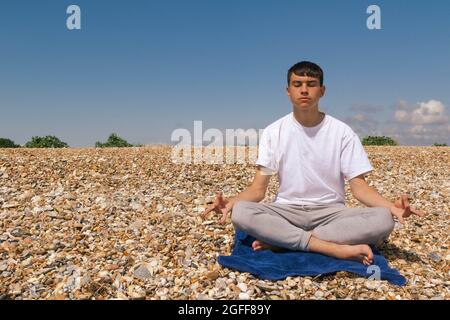 Un adolescent de race blanche méditant sur une plage pierreuse avec ses mains en position Shuni Banque D'Images