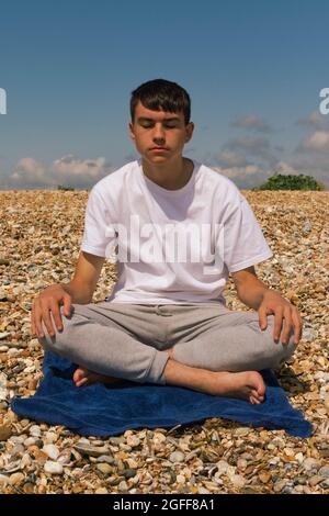 Un adolescent du Caucase méditant sur une plage pierreuse avec ses mains reposant sur ses genoux Banque D'Images