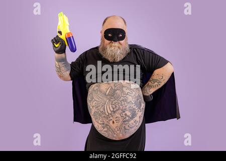 Homme barbu avec grand abdomen tatoué en costume de carnaval tient le blaster jouet sur fond violet Banque D'Images