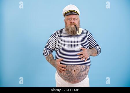 L'homme obèse d'âge moyen en costume de marin tient les mains sur le ventre sur fond bleu clair Banque D'Images