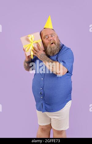 Drôle d'homme avec le surpoids met l'oreille à boîte cadeau posant sur fond violet en studio Banque D'Images