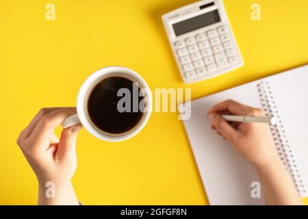 gros plan à portée de main tenant un crayon écrivant sur le carnet à côté de la calculatrice sur fond jaune - planification de bureau et d'affaires ou objectifs pause café concep Banque D'Images
