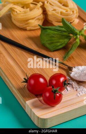 Image de tomates cerises au premier plan sur une table en bambou avec de la farine, du basilic et des pâtes en arrière-plan, le tout sur un fond vert Banque D'Images