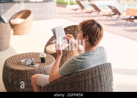 jeune homme allongé sur la chaise longue et lisant un livre près de la piscine Banque D'Images