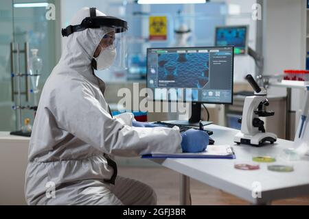 Chimiste chercheur écrivant l'expertise du virus sur le presse-papiers développement du vaccin de prévention du coronavirus pendant l'enquête microbiologique. Scientifique en costume médical de protection travaillant dans un laboratoire hospitalier Banque D'Images