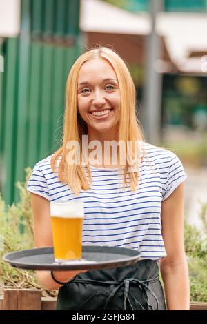 Une serveuse jeune femme sympathique et heureuse propose un verre de bière fraîche sur un plateau avec un sourire éclatant dans un restaurant ou un pub en plein air Banque D'Images