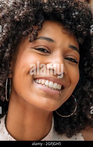 Portrait d'une belle femme noire souriante et sympathique avec des cheveux bouclés et des piercings de nez et de lèvre regardant l'appareil photo avec des yeux riant dans un gros plan Banque D'Images