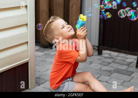 Portrait de mignon petit garçon d'enfant de lien profiter d'avoir amusant jouer bulles de savon soufflant siting à la cour à la maison à l'extérieur sur la lumière chaude d'été contre la maison Banque D'Images