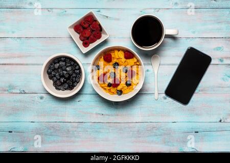 Petit déjeuner de travail sur une surface en bois de céréales cornflakes avec framboises et bleuets dans un bol blanc avec cuillère blanche, smartphone et café Banque D'Images