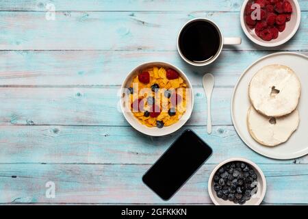 Petit déjeuner de travail sur une surface en bois de céréales cornflakes avec framboises et bleuets dans un bol blanc avec cuillère blanche, smartphone et café Banque D'Images