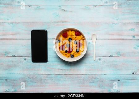 Petit déjeuner de travail sur une surface en bois de céréales de céréales de cornflakes avec framboises et bleuets dans un bol blanc avec une cuillère blanche et un smartphone. Banque D'Images