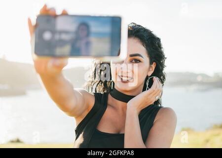 Une jeune femme heureuse portant une robe noire prenant un smartphone devant son visage et prenant un selfie Banque D'Images