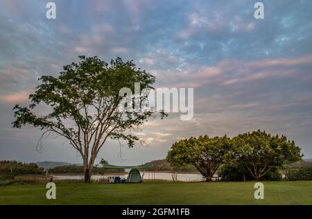 Vue d'une tente de camping avec auvent perché sur l'herbe sous de beaux grands arbres avec des branches au bord du lac du camping avec chaîne de montagnes à l'arrière Banque D'Images