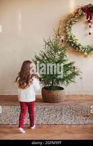 Jolie petite fille sur le tapis sous par arbre de noël décoré pendant la saison des fêtes d'hiver, portant un chandail blanc boutonné avec pantalon à carreaux Banque D'Images