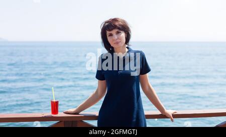 Une brunette souriante avec un verre de jus se trouve sur la jetée au bord de la mer. Banque D'Images