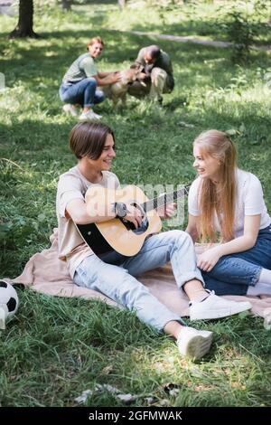 Des adolescents souriants jouant de la guitare acoustique à proximité d'amis flous avec un chien sur la pelouse Banque D'Images