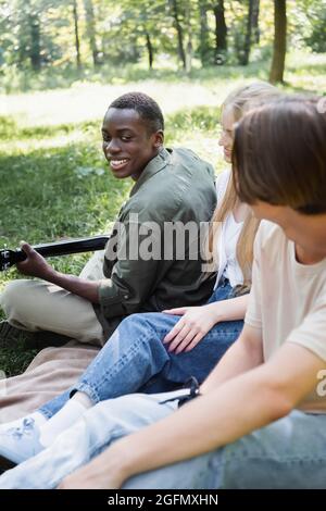 Un adolescent afro-américain souriant jouant de la guitare acoustique près d'amis flous dans le parc Banque D'Images