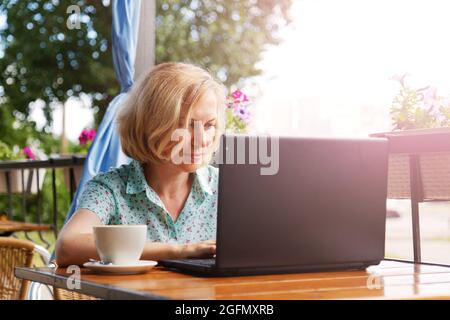 Seule femme assise à la table avec un ordinateur portable et une tasse de café sur la véranda ouverte d'été. Mise au point peu profonde. Concept de travail hors bureau. Banque D'Images