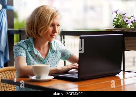 Seule femme assise à la table avec un ordinateur portable et une tasse de café sur la véranda ouverte d'été. Mise au point peu profonde. Concept de travail hors bureau. Banque D'Images