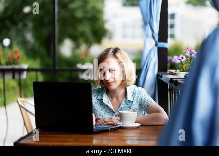 Seule femme assise à la table avec un ordinateur portable et une tasse de café sur la véranda ouverte d'été. Mise au point peu profonde. Concept de travail hors bureau. Banque D'Images