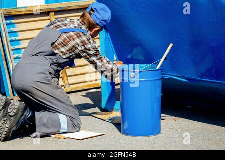 Un vieil homme dans une combinaison de travail peint une poubelle ou un conteneur dans la rue avec un pinceau. Travail à temps partiel pour un pensionné. Toil. Banque D'Images