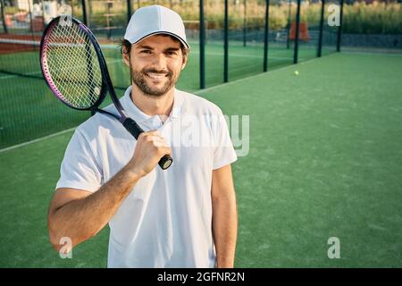 Portrait d'un beau joueur de tennis avec une raquette à la main debout sur un court de tennis portant un uniforme blanc Banque D'Images