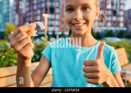 Une petite fille positive tenant la dernière génération d'aide auditive dans sa main dans la rue, en plein air . Traitement de la surdité chez les enfants et solution auditive Banque D'Images