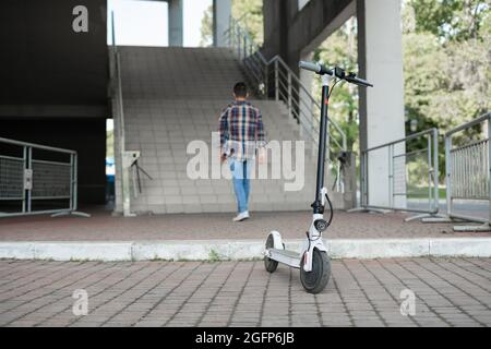 L'homme place le scooter électrique à l'extérieur du centre commercial et part. Vue de l'arrière sans visage. Mode de vie technologique écologique. Banque D'Images