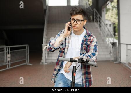 Homme portant des lunettes homme d'affaires avec un scooter électrique debout devant un bâtiment d'affaires moderne parlant au téléphone. Banque D'Images