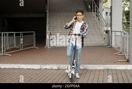 Homme portant des lunettes homme d'affaires avec un scooter électrique debout devant un bâtiment d'affaires moderne parlant au téléphone. Banque D'Images