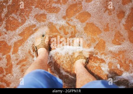 Pieds pour hommes dans des sandales sur la plage. Homme en Jean short se tient et regarde ses pieds dans le sable qui se lave par l'océan. L'été. Reposez-vous à l'hôtel. Banque D'Images