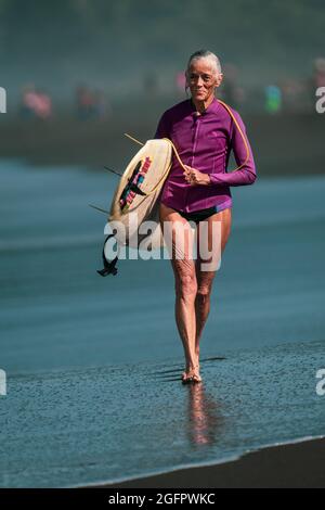 Playa Hermosa, Guanacaste, Costa Rica - 07.26.2020: Une femme de surfeur senior avec des cheveux gris marche joyeusement sur la plage avec une planche de surf. Banque D'Images