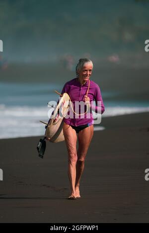 Playa Hermosa, Guanacaste, Costa Rica - 07.26.2020: Une femme de surfeur senior avec des cheveux gris marche joyeusement sur la plage avec une planche de surf. Banque D'Images