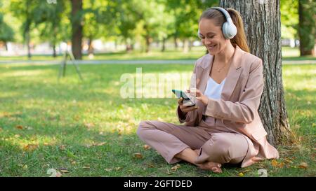 Photo d'une femme de 40 ans souriante, portant un casque avec un smartphone. Elle écoute sa musique préférée tout en étant assise sur l'herbe dans le p Banque D'Images