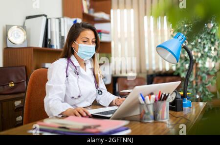 Femme hispanique médecin en masque facial assis au bureau Banque D'Images