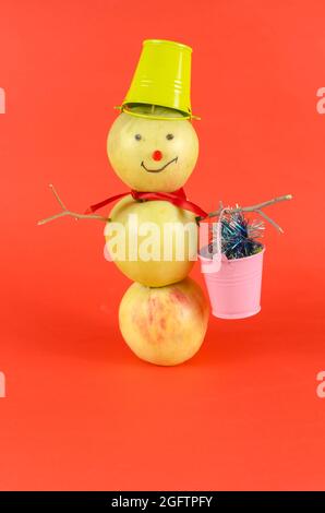 Bonhomme de neige Apple avec un seau vert sur sa tête debout sur un fond rouge. Bonhomme de neige créatif de trois fruits mûrs avec un seau rose plein de vacances g Banque D'Images