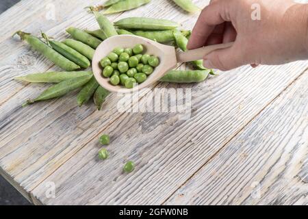 La main d'une femme tient une cuillère en bois avec des pois verts sur une table en bois clair. Banque D'Images