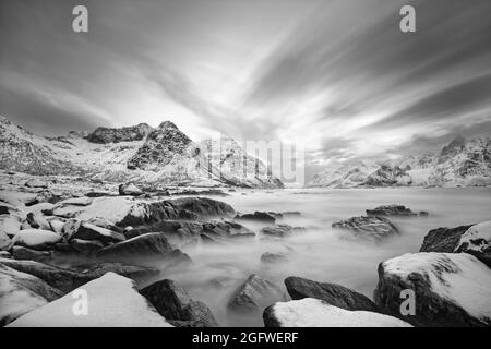 Image en noir et blanc d'un paysage côtier en hiver avec mouvement d'eau entre de grandes pierres avec de la neige sur elles, en arrière-plan une chaîne de montagnes Banque D'Images