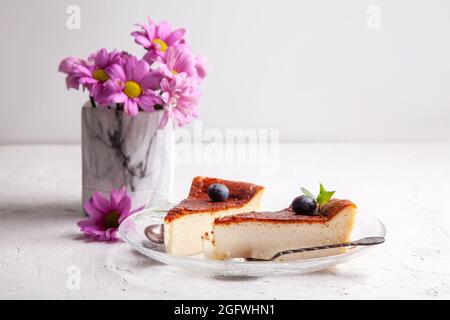 Des tranches de cheesecake basque maison ont brûlé sur une assiette avec des bleuets et des feuilles de menthe sur fond clair avec des fleurs dans un vase Banque D'Images