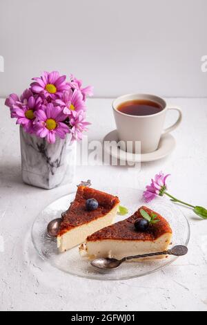 Tranches de cheesecake basque maison brûlé sur plaque avec bleuets et feuilles de menthe, tasse de thé noir sur fond clair avec fleurs dans vase Banque D'Images
