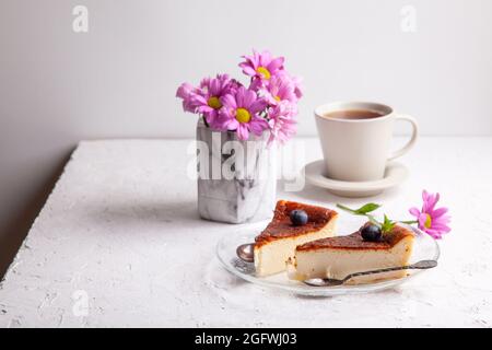 Tranches de cheesecake basque maison brûlé sur plaque avec bleuets et feuilles de menthe, tasse de thé noir sur fond clair avec fleurs dans vase Banque D'Images