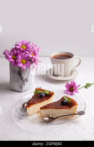Tranches de cheesecake basque maison brûlé sur plaque avec bleuets et feuilles de menthe, tasse de thé noir sur fond clair avec fleurs dans vase Banque D'Images