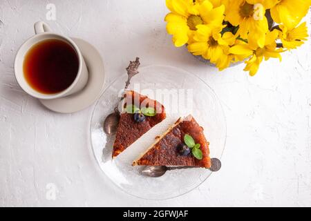 Tranches de cheesecake basque maison brûlé sur plaque avec bleuets et feuilles de menthe, tasse de thé noir sur fond clair avec fleurs dans vase Banque D'Images